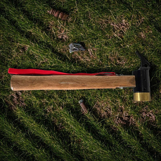 Camping mallet hammer with wooden handle, black metal head, brass peg puller, red lanyard, on green grass with scattered tent pegs