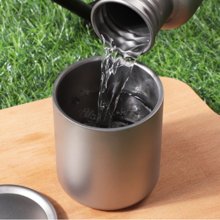 Water is poured from the kettle into a Boertala titanium cup placed on a wooden surface, with green grass as the background.