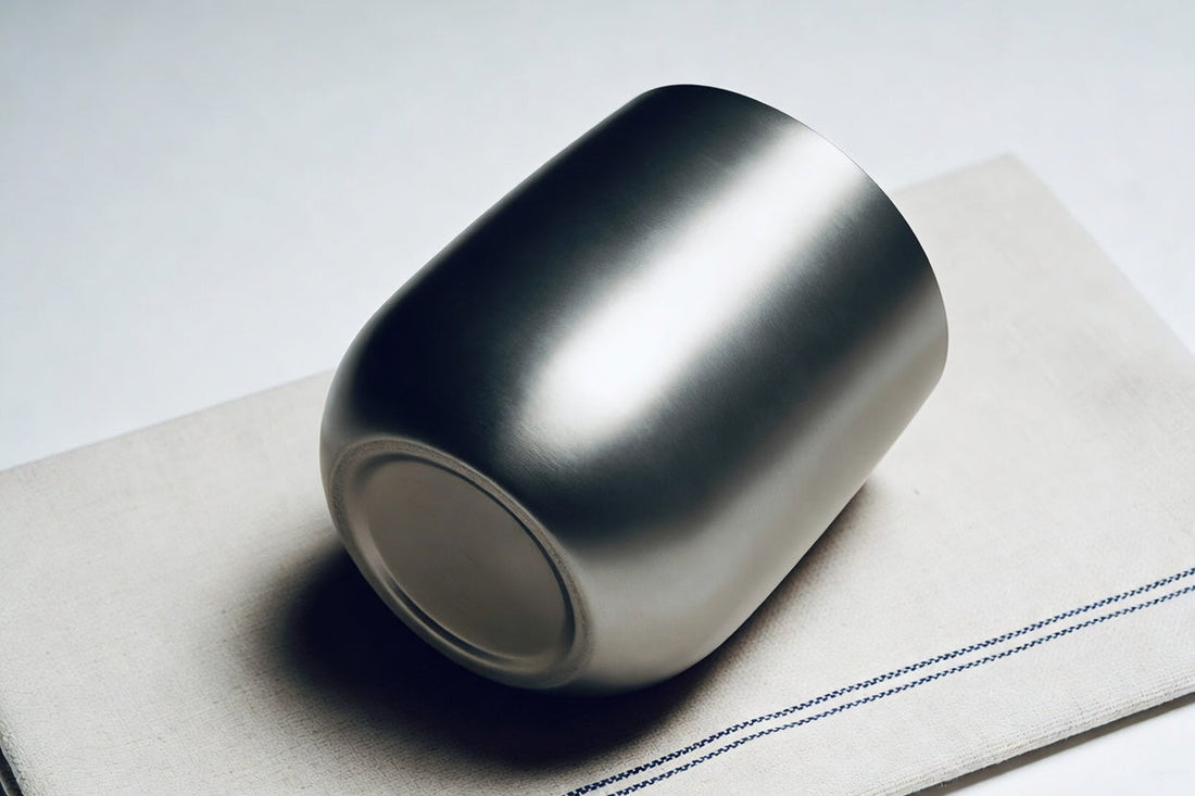 Silver titanium metal water cup lying on its side on a white linen napkin with blue stripes, smooth surface with reflective highlights, white inner wall visible at the base.