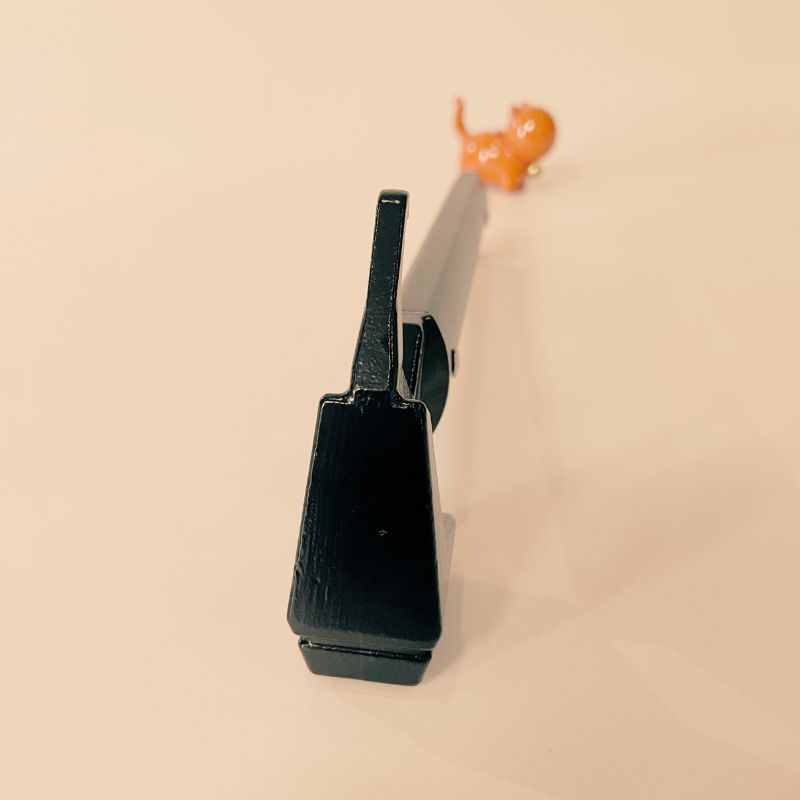 Black gun-shaped object on a beige background with an orange toy figure in the corner.