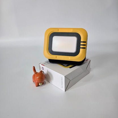 Yellow and black floodlight on a white box with an orange cat figurine next to it on a light gray background