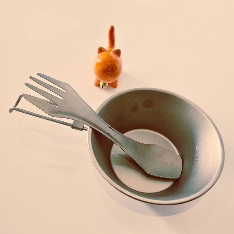 Set of cutlery including a fork, knife, and spoon in a gray bowl with a small orange cat figurine on a beige background.