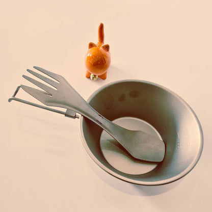 Set of cutlery including a fork, knife, and spoon in a gray bowl with a small orange cat figurine on a beige background.