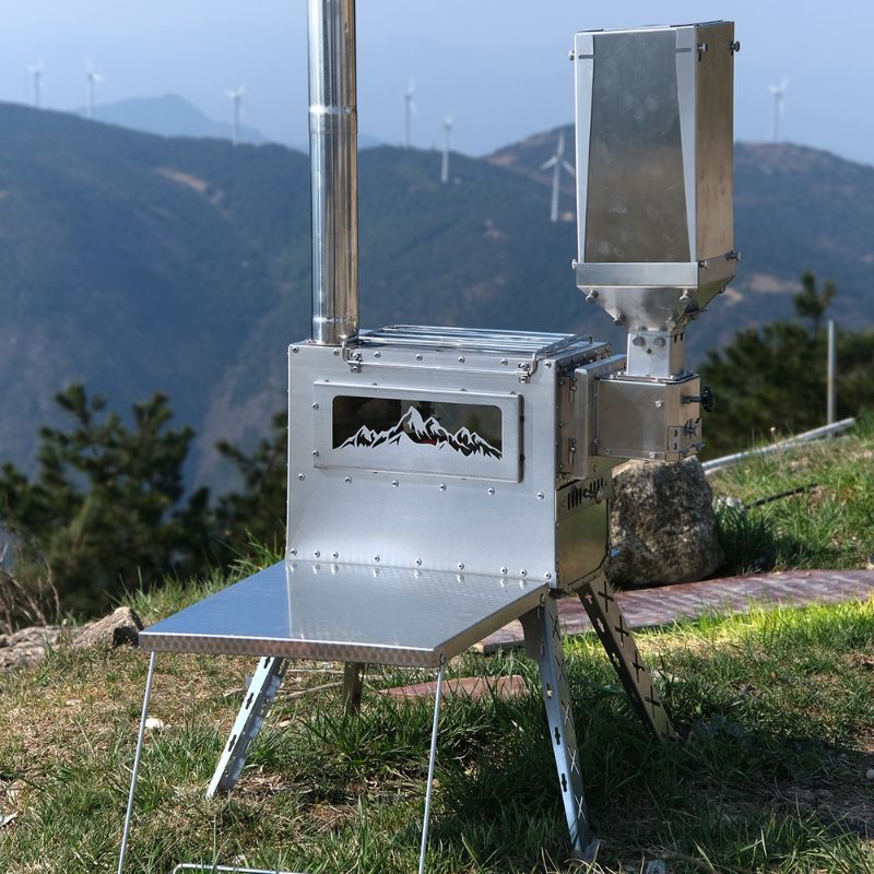 Metallic camping stove with mountain design on a grassy area with wind turbines in the background