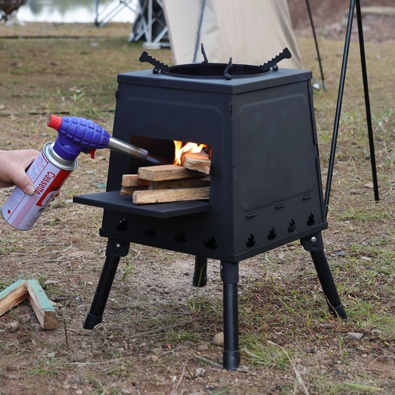 Person using a butane torch to light wood in a black outdoor stove in a natural setting.