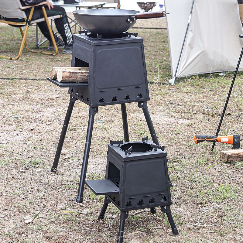 Black cast iron stove on legs with a frying pan over it, set up outdoors with a tent in the background.