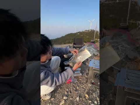 Person unboxing and assembling a premium stainless steel outdoor wood burning stove with built-in oven in a scenic camping location with wind turbines