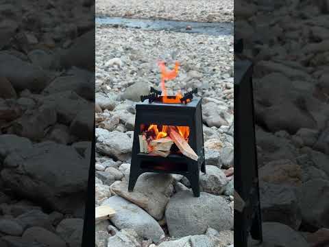 Black portable foldable carbon steel tower camping wood stove burning brightly on rocky riverbank outdoors