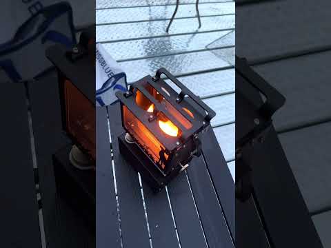 Lit kerosene lantern stove with glowing flame and deer silhouette creating cozy camping atmosphere on wooden table outdoors