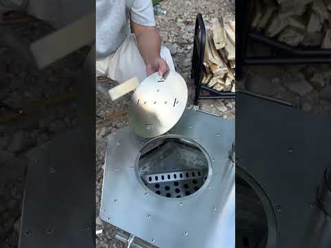 Person adding firewood to a stainless steel hexagonal tower wood stove outdoors for camping cooking demonstration
