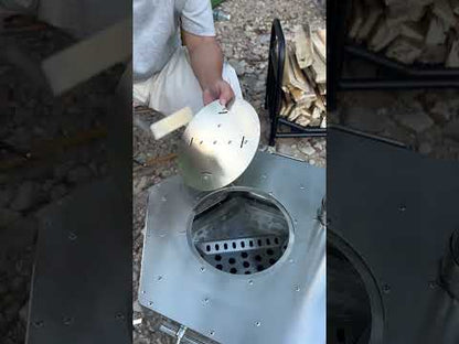 Person adding firewood to a stainless steel hexagonal tower wood stove outdoors for camping cooking demonstration